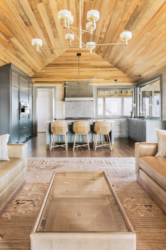 Golden Hues: In the open-plan living/kitchen space, the ceiling of honey-colored, hand-placed sunken cypress planks contrasts beautifully with the custom cabinetry painted in “Down Pipe” by Farrow & Ball. A vibrant tile backsplash, clad in “Hip Herringbone Detroit Blues” by Artistic Tile, sits under a custom exhaust hood made by Handcrafted Metal. Three McGuire rattan stools at the island offer occupants a view through the sliding window pass-through to the dining porch.