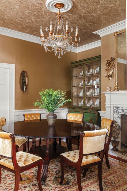 A Warm REception: Halsey & Spruce dupioni silk-covered walls pair with the Twigs “Villa Foscari”-papered ceiling in the dining room to create a feeling of grandeur in the small room. Overlooked by the George III Japanese cabinet in green and gold, a custom, round mahogany table with Chesterfield dining chairs upholstered in gold velvet provide enough space for the Griffin family to congregate.