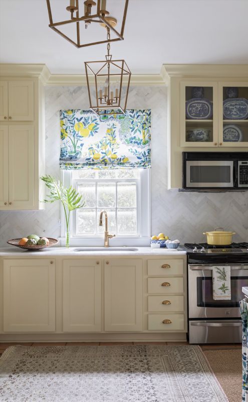 Bright Ideas: A colorful Schumacher fabric, hanging above the window and concealing a washer and dryer (above), makes a bold statement in this traditional cottage kitchen. Soft yellow Farrow & Ball “Pale Hound” paint paired with brass hardware on the cabinets creates a vintage feel, while a white marble backsplash in a herringbone pattern and a giclée print by Hayley Mitchell add touches of modern luxury.