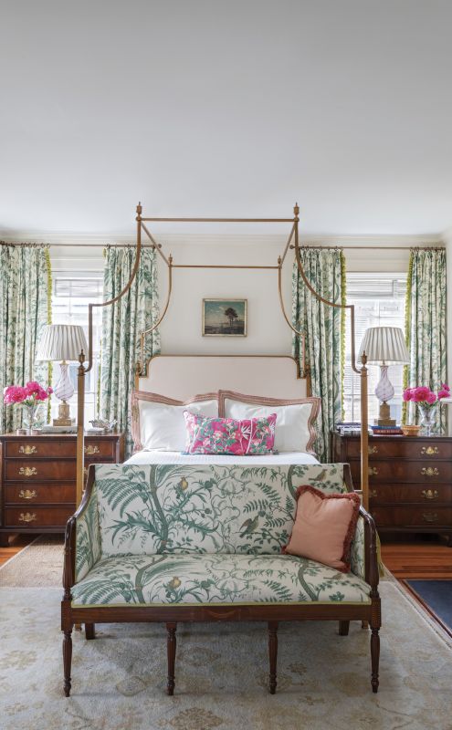FIT FOR A QUEEN: A quieter color palette in the primary bedroom creates a soothing, luxurious space. The delicate design of the Brunschwig & Fils “Bird & Thistle” toile fabric for the curtains and the antique Sheraton settee, the brass four-poster bed, and vintage Murano glass lamps combine to produce a light and feminine feel. The theme continues in the bath with a white Carrara marble tiled tub and vanity highlighted with accents of Kelly green.
