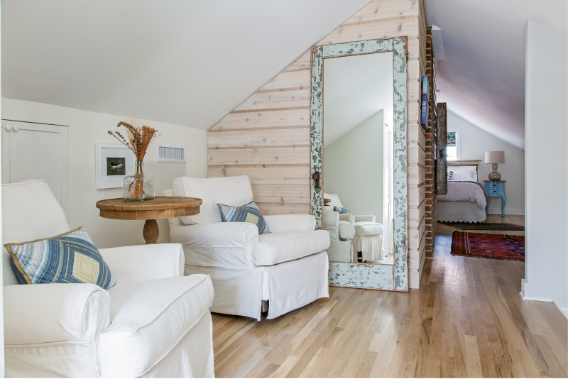 ABOVE & BEYOND: The second floor houses a relaxing master suite. Dimmable recessed lighting, comfy upholstered chairs from Celadon, and textural elements—cedar planks, a Past to Present Décor mirror made from salvaged wood, and exposed brick—amp up the space’s homey vibe. Pops of color via the Roman shades made with fabric from GDC Home, the teal bedside table from Nadeau, and vintage rugs enliven the design’s neutral palette.