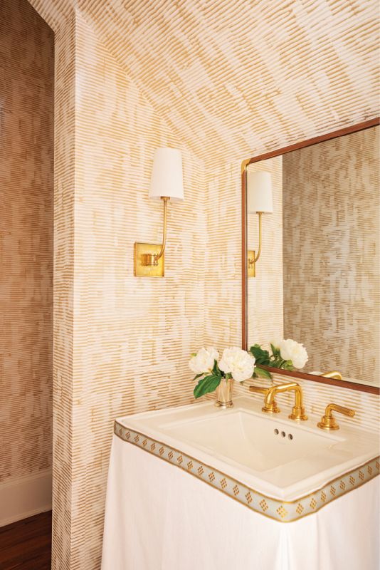 A textured gold wallpaper from Romo in the nearby powder room adds pizazz, with its metallic tones picked up in the “Venini” gold sconce from Circa Lighting, the faucet, and the custom white linen sink skirt with a Samuel & Sons trim.