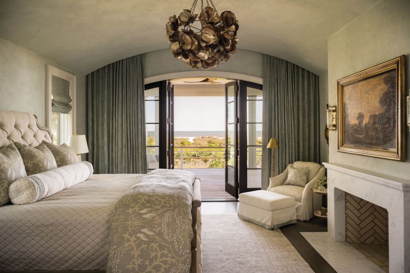 The primary bedroom features a curved plaster ceiling and walls, enhancing the warmth of the cozy space. Susan selected an oil painting of the English countryside from her Gainesville home for above the fireplace, opting to hide the TV in a nearby armoire. “I love that piece. I’d rather look at it than a TV,” she says. Deep green drapes and patterned bedding from Lewis & Wood, Imogen Heath, and Cowtan & Tout add interest.