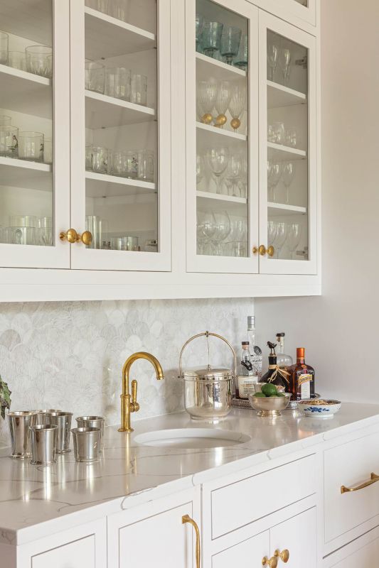 A sculptural wet bar pairs quartzite countertops with a mosaic backsplash. Glass-front cabinets display the family’s collection of stemware, while brass hardware adds warmth.