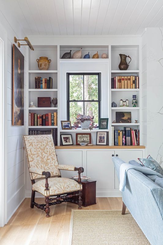 Tucked near the bookshelves, an antique chair in crewel fabric invites a quiet read. Above and around it, books, framed photographs, and personal mementos form the “last layer” designer Melissa Ervin loves: that final, lived-in feel of family history.