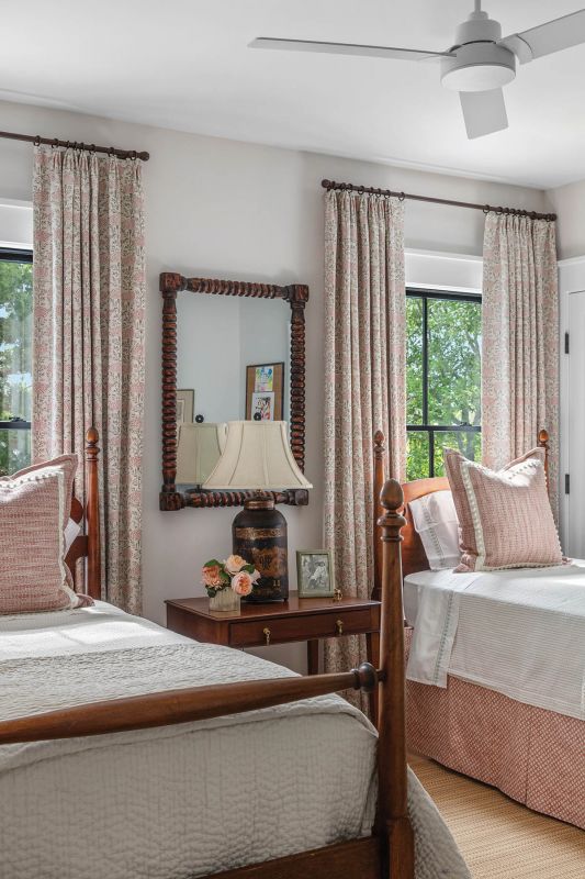Antique twin beds, an inherited side table, and a tea caddy lamp create a soft, pink-tinged nest perfectly suited to two visiting granddaughters. Lisa Fine “Samode” fabric in “Poppy” forms the curtains, and pillows trimmed with Samuel & Sons pom-poms add a playful touch.
