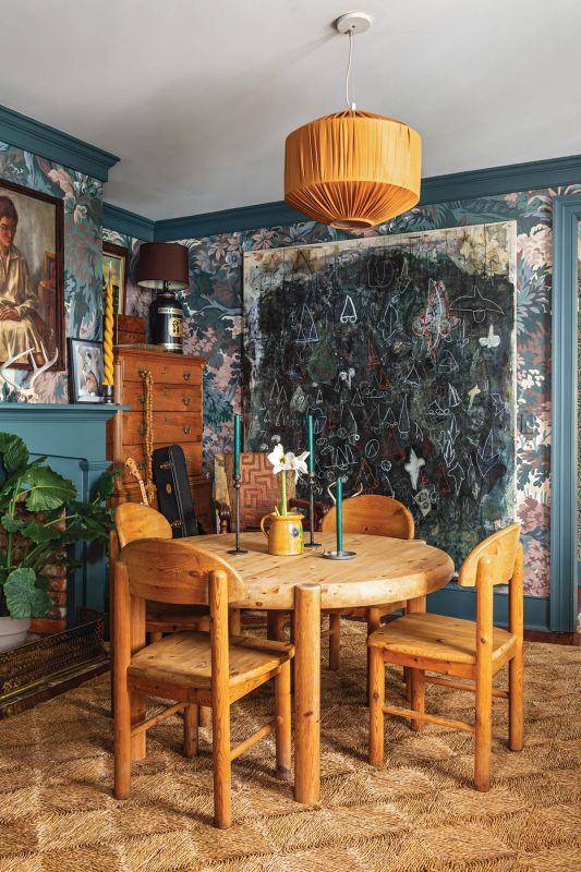 Moody Hues: The dining room forms the dramatic heart of the home, wrapped in the fantastical House of Hackney “Arborea” wallpaper. A large abstract painting by Doug Higgins anchors the space, its dark palette offset by the light pine of a 1970s Rainer Daumiller dining set, which is topped by a color-matched Pierce & Ward pleated raw silk pendant. A Rush House braided sea-grass rug adds rustic texture underfoot.