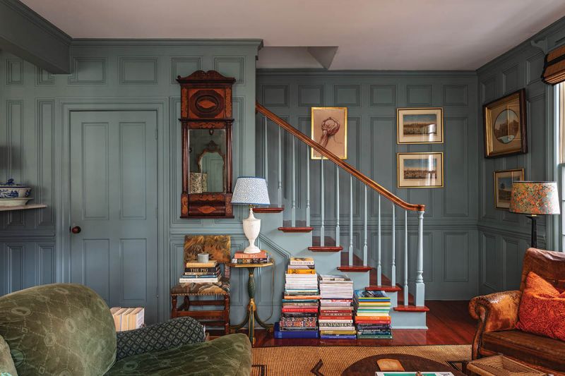 An Artful Retreat: The colorful den at the rear of the home, part of a later addition, features paneled walls painted in Farrow & Ball “Oval Room Blue” and adorned with art, including local artist Jill Hooper’s Drawing of Headdress, alongside antique pieces.