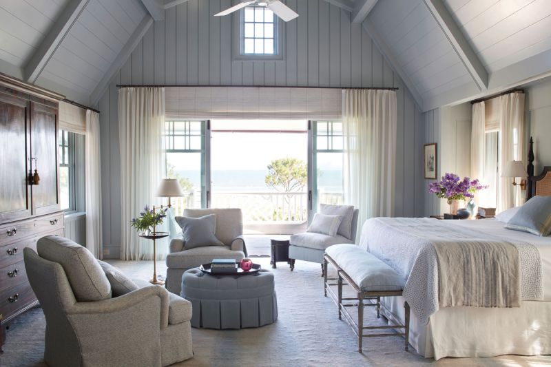 Sleeping bEauty: In the primary bedroom, decorative woodwork painted in Farrow & Ball’s “Light Blue 22” evokes the deep blue ocean beyond the picture frame window. Sheer drapes allow the sunlight to stream through and comfy club chairs from TCS Designs pair with an antique armoire and bed for a touch of relaxed luxury. An ottoman wrapped in Rogers & Goffigon fabric and a bench from Suzanne Rheinstein’s Hollyhock complete the look.